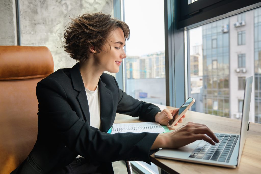 Young working woman in office sitting in front of computer and using smartphone wearing suit