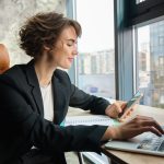 Young working woman in office sitting in front of computer and using smartphone wearing suit