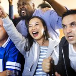 Diverse global sports fans from different countries celebrating an exciting moment while watching multilingual broadcast coverage