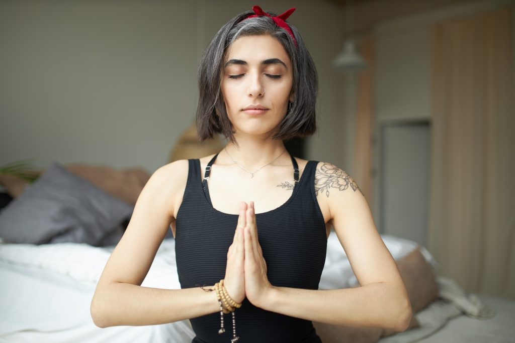 Portrait of mindful attractive young female yogi with gray hair and tattoo sitting in lotus pose keeping hands pressed together on her chest