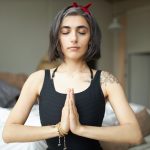 Portrait of mindful attractive young female yogi with gray hair and tattoo sitting in lotus pose keeping hands pressed together on her chest